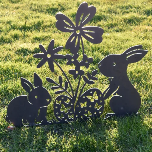 Metalfigur i cortenstål med to kaniner, blomster og påskeæg – dekorativ havefigur "Forårsglæde" fra Onlinesmeden, fotograferet i sollys på grøn græsplæne.